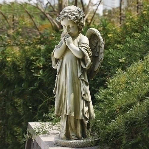 Praying angel child garden statue outdoors with shrubs behind it.