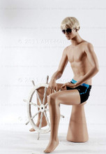 In this full body side view photo, wearing a short blond wig / hairpiece and a black swim suit,  mannequin Roger sits upright with his legs apart and his hands in his lap.  Mannequin Roger can be displayed with or without a wig / hairpiece.  Pedestal included (sunglasses not).