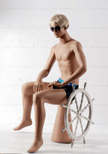 In this full body  side view photo, wearing a short blond wig / hairpiece and a black swim suit,  mannequin Roger sits upright with his legs apart and his hands in his lap.  Mannequin Roger can be displayed with or without a wig / hairpiece.  Pedestal included (sunglasses not).